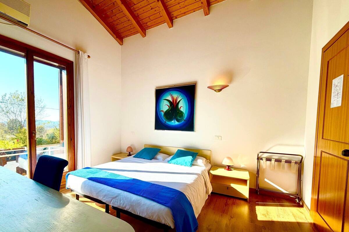 Une chambre à coucher avec un lit, des tables de chevet, une lampe, un radiateur et une fenêtre donnant sur un beau paysage.
