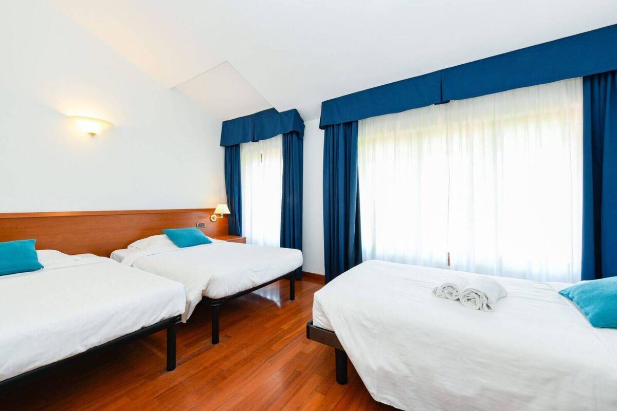 Une chambre d’hôtel avec trois lits simples, un plancher en bois, une table de chevet avec une lampe et de grandes fenêtres avec des rideaux bleus.