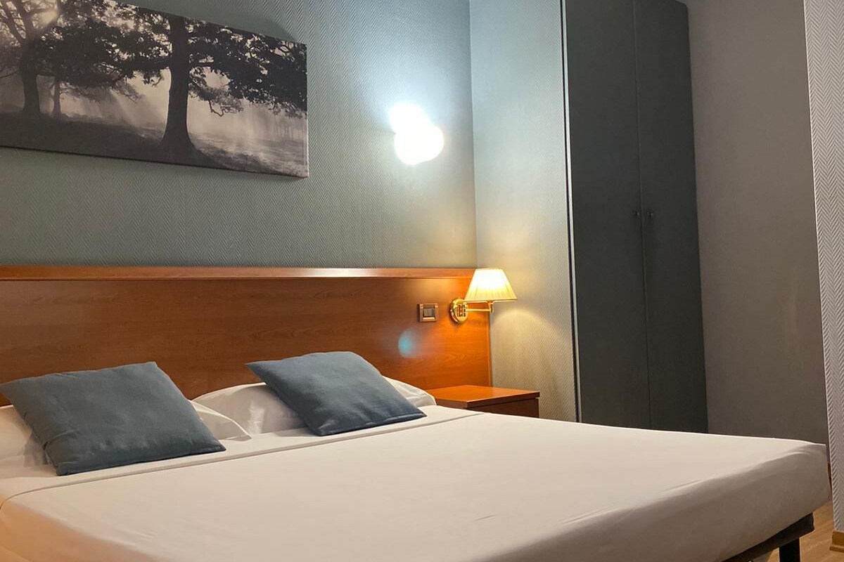 Een hotelkamer met een groot bed, een nachtkastje met lamp, een aan de muur hangend schilderij en een kledingkast.