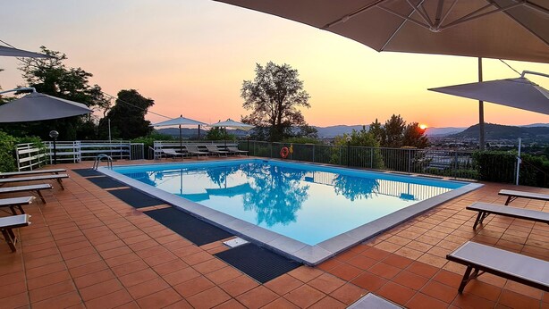 A swimming pool with lounge chairs and umbrellas, surrounded by a tiled deck and a view of the sunset over a hilly landscape. - Hotel New Genziana (Altavilla Vicentina)