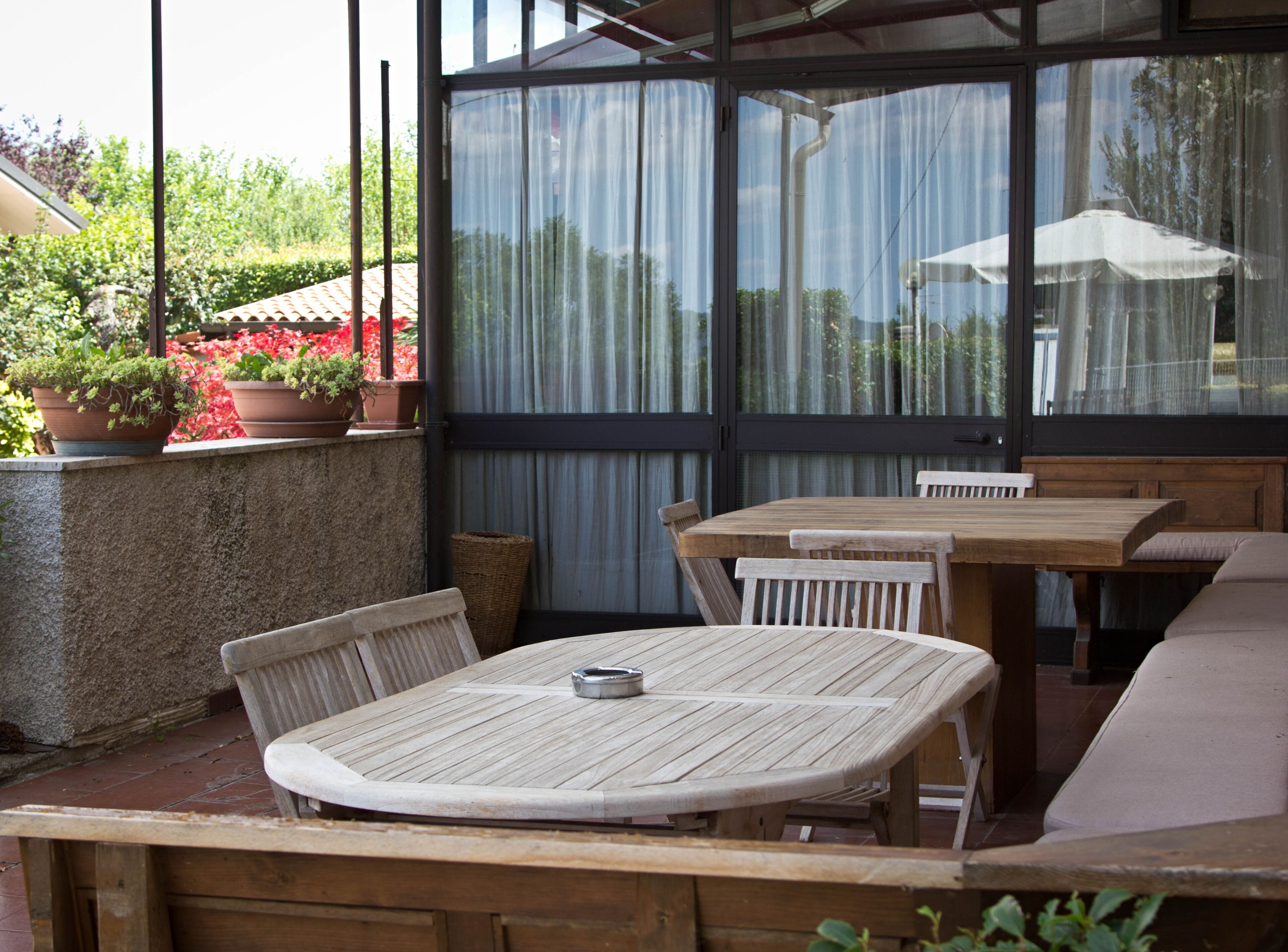 Un patio al aire libre con mesas y sillas de madera, plantas en macetas y vistas a través de un ventanal.