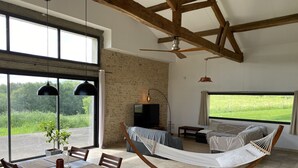A spacious living room with a large window, a hammock, a dining area with a table and chairs, and a television. - Converted barn/gergergerie in the countryside. (Castelner)