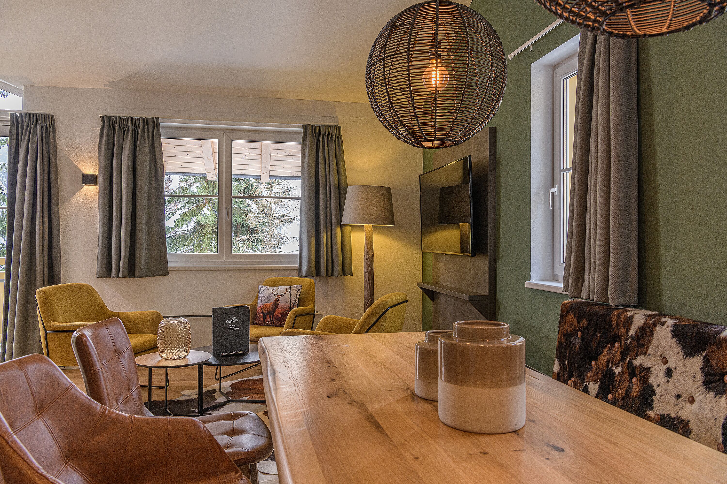 Un salon moderne avec une table à manger en bois, des fauteuils en cuir et un canapé au motif original. On y trouve deux lampes, un téléviseur et une fenêtre avec des rideaux.