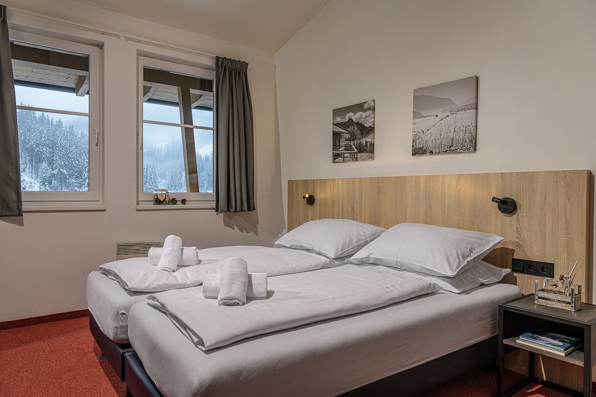 Ein Doppelbett mit weißer Bettwäsche und Handtüchern, ein Nachttisch mit Lampe sowie zwei gerahmte Bilder an der Wand.