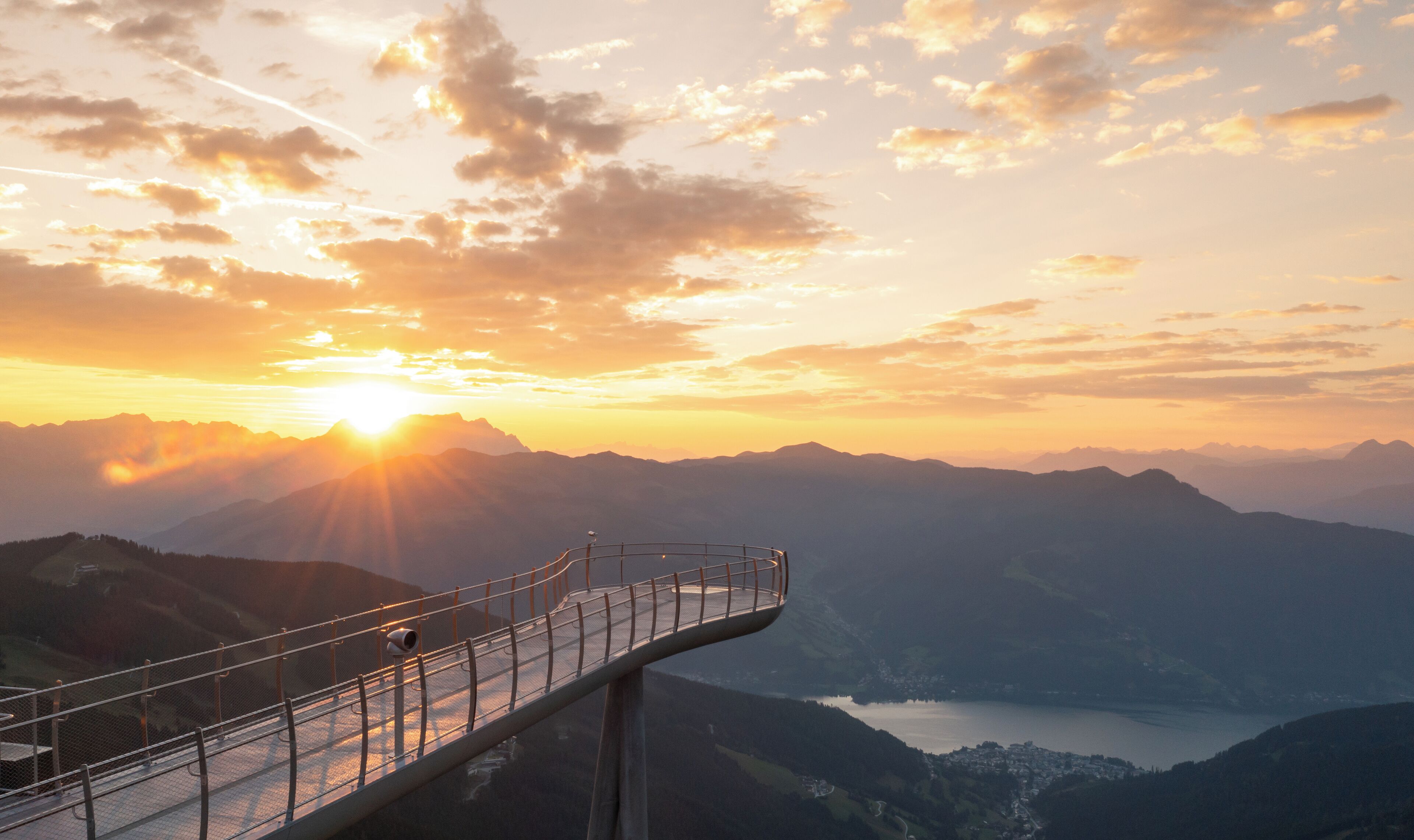 A scenic overlook with a metal railing, overlooking a valley and a lake during sunset.