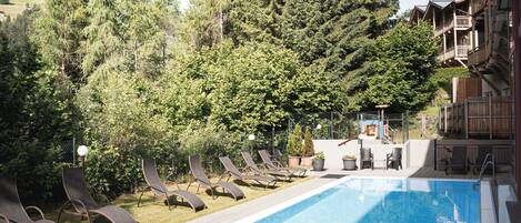 A swimming pool with lounge chairs and a wooden deck, surrounded by trees and a building with balconies.
