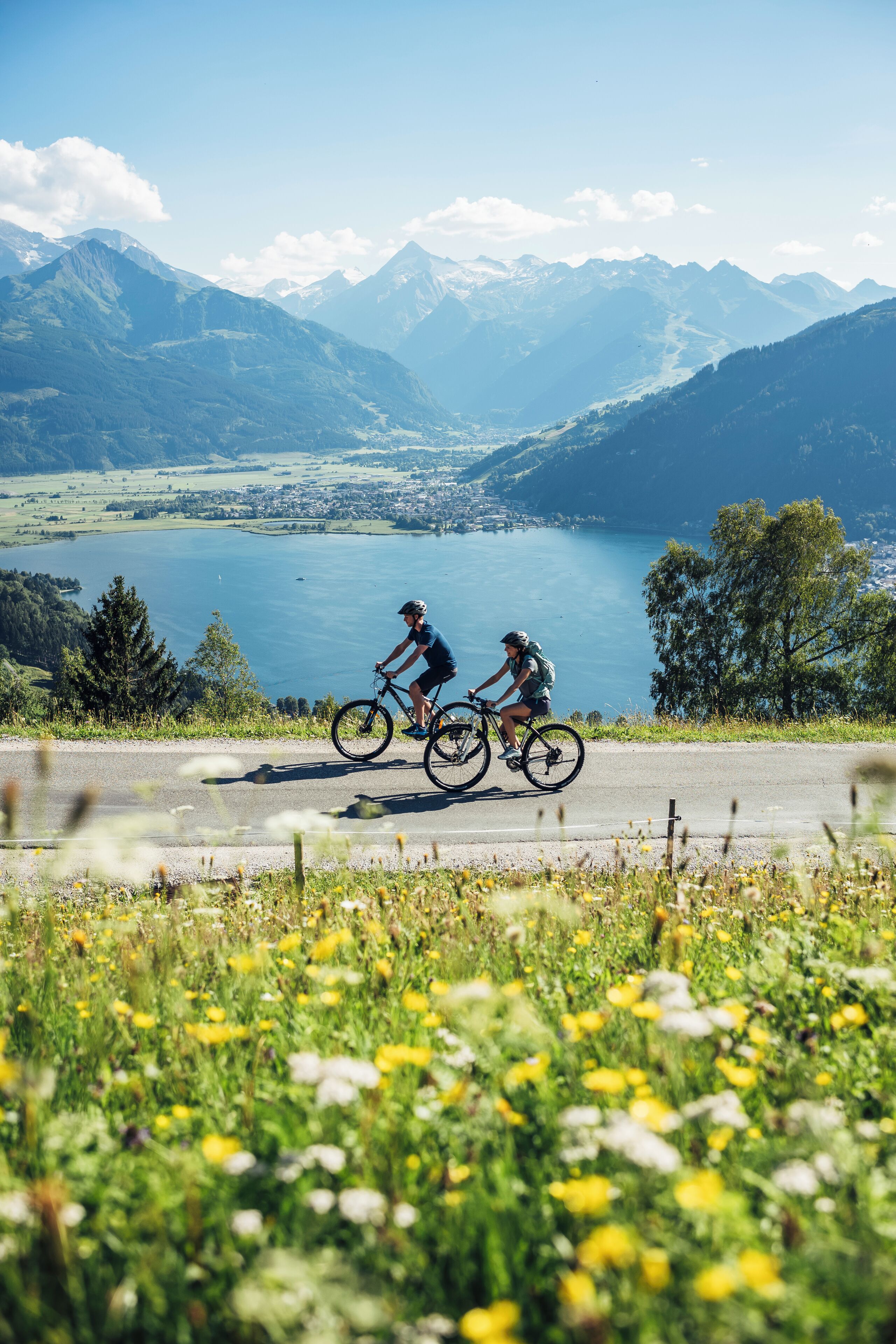 Two cyclists on a road with a mountainous landscape and a lake in the background.
