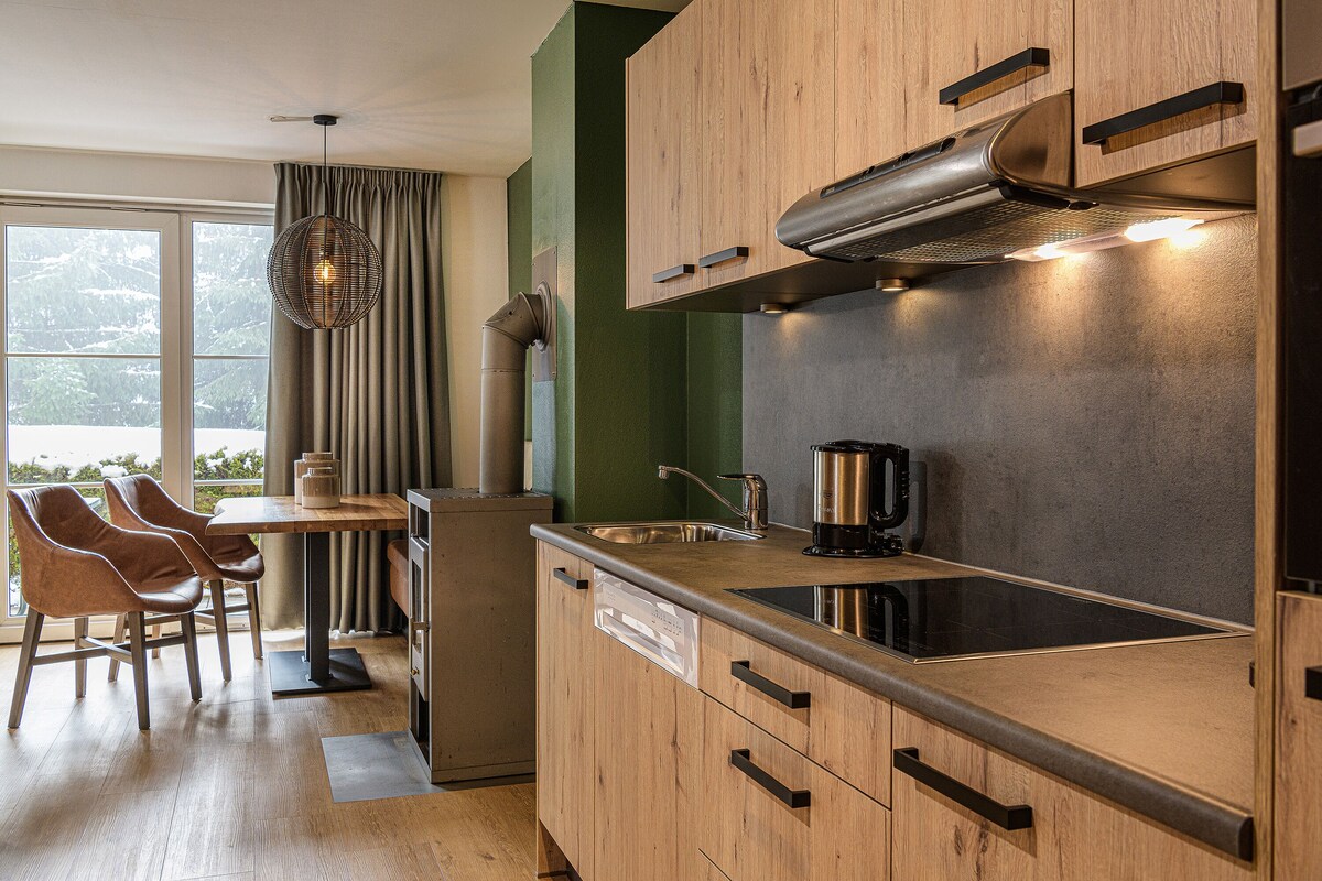 A modern kitchen with wooden cabinets, a stainless steel sink, and a built-in stove. There is a dining area with chairs and a table.