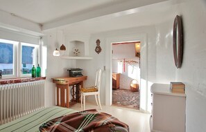 Una habitación con una cama, un escritorio con una máquina de escribir, una silla, un radiador y una ventana con botellas.