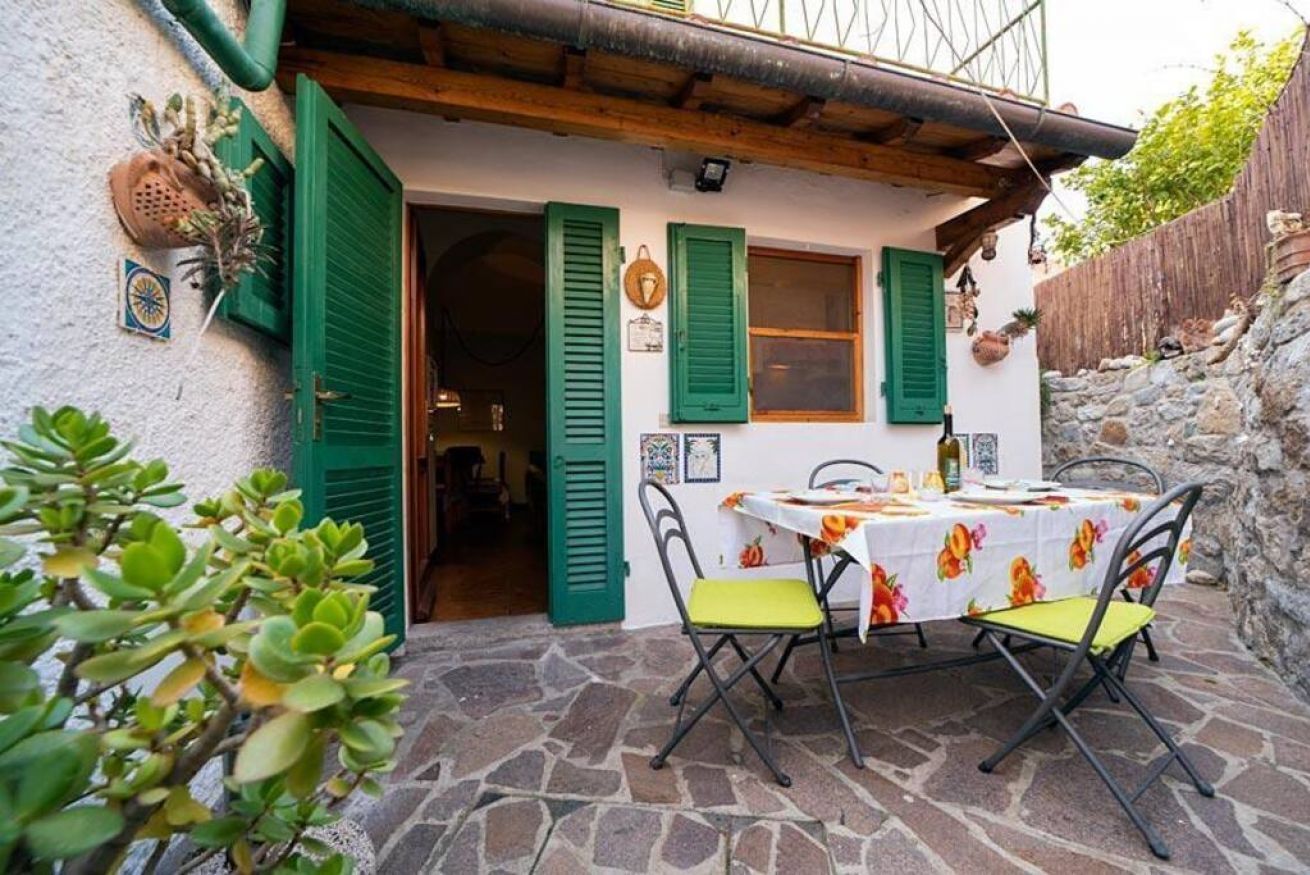 A stone-floored patio with a table and chairs, green shutters, and a rustic building exterior.