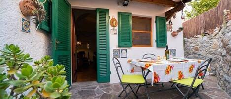 A stone-floored patio with a table and chairs, green shutters, and a rustic building exterior.
