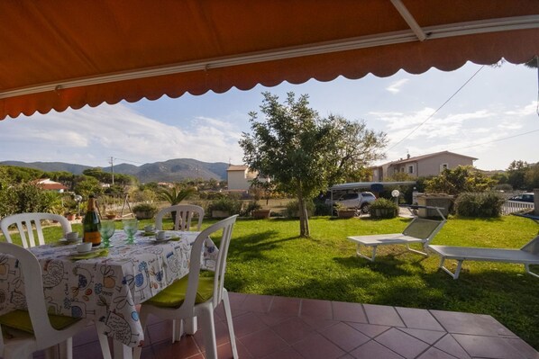 Une terrasse extérieure avec une table dressée pour le repas, entourée de chaises, et une pelouse avec un arbre et une voiture en arrière-plan.