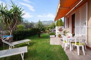 A patio with a table and chairs, a small garden with a palm tree, and a view of the hills. - Margherita 1 apartment (Marina di Campo)