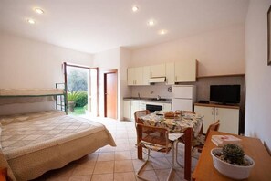 A bedroom with a bed, a dining table with chairs, a small kitchen area with cabinets and a television, and a door leading to a balcony.