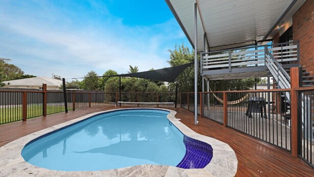 A swimming pool with a wooden deck, a covered seating area, and a brick wall in the background.