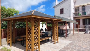 A wooden gazebo with benches and a table, a stone patio, and a white building with a balcony in the background. - Aricipelgo - Ponza apartment (Marina di Campo)