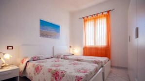 Une chambre avec deux lits, une table de chevet, une lampe, un tableau représentant une plage et une fenêtre avec des rideaux orange.