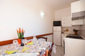 A small kitchen with a refrigerator, stove, and microwave. There is a dining table with chairs and a tablecloth.