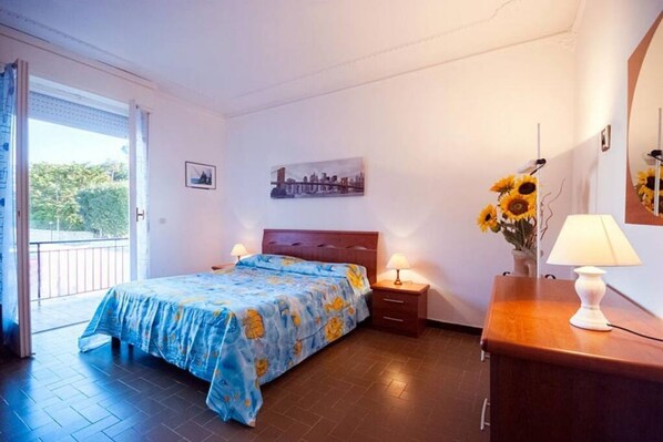 Ein Schlafzimmer mit Bett, Nachttisch, Lampe, einer Vase mit Sonnenblumen, einem Bild an der Wand und Blick durch eine Tür.