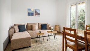 Een woonkamer met een bank, twee salontafels, een houten eettafel en stoelen. Twee ingelijste foto’s hangen aan de muur en er is een raam met gordijnen.