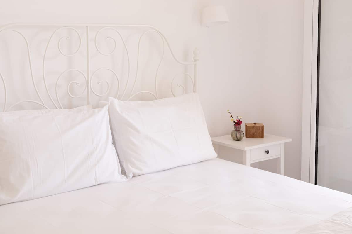A white metal bed frame with a simple headboard, a white bedside table with a drawer, and a vase with flowers.