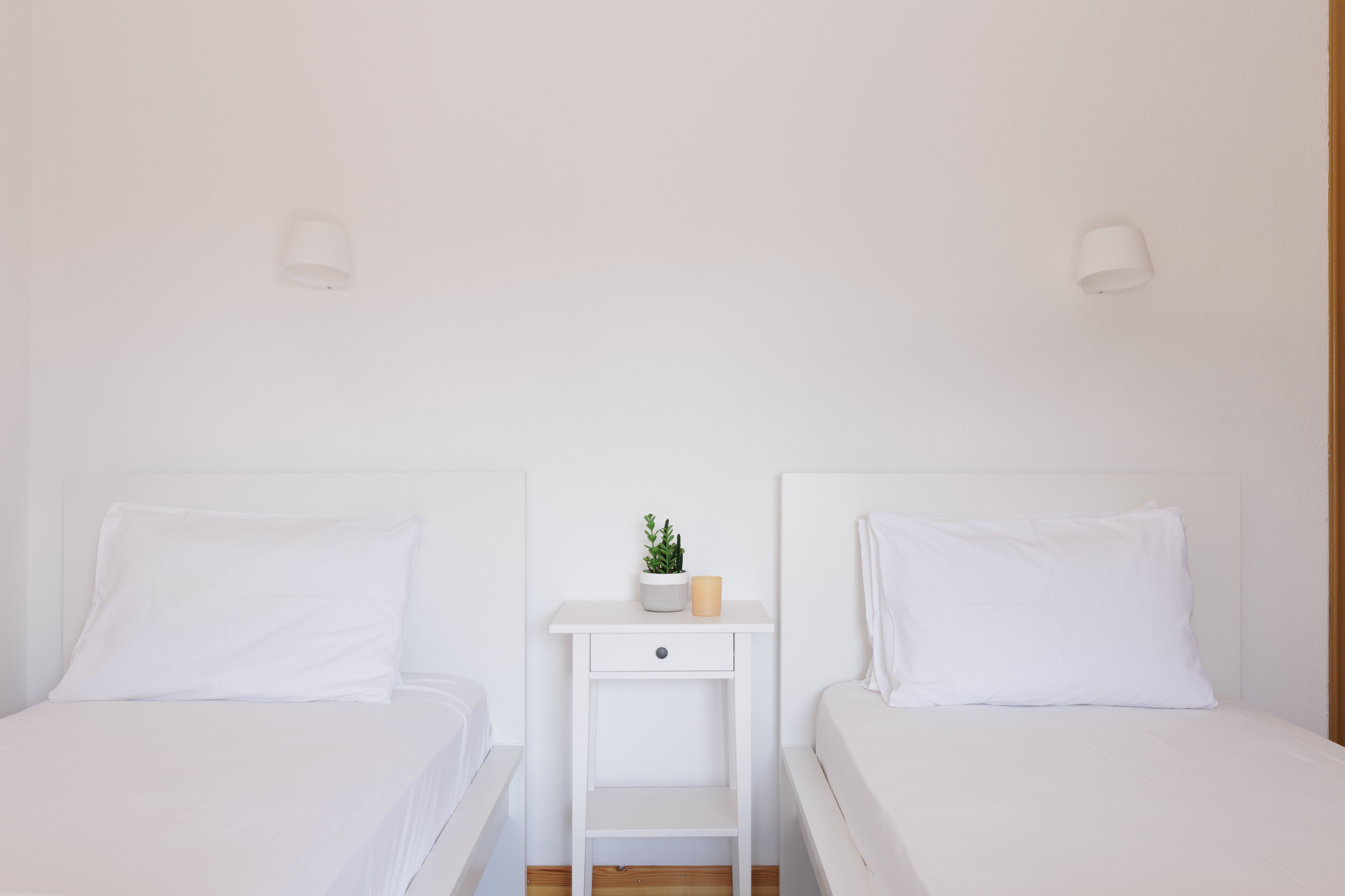 A clean, minimalist bedroom with two white beds, a small bedside table with a plant, and two wall-mounted lamps.