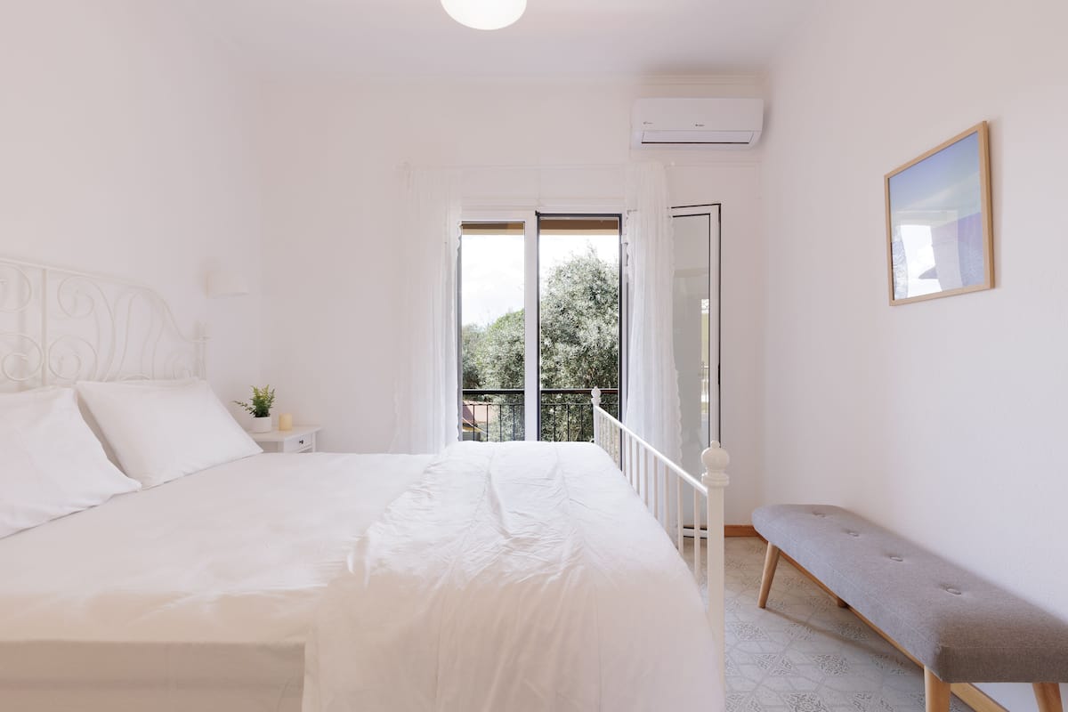 A bedroom with a bed, a bench, a nightstand, a window, and an air conditioning unit.