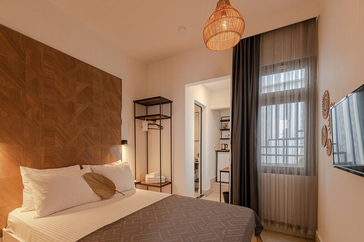 A modern bedroom with a large bed, a wooden headboard, a bedside table, a hanging light fixture, and a view of a hallway and bathroom.