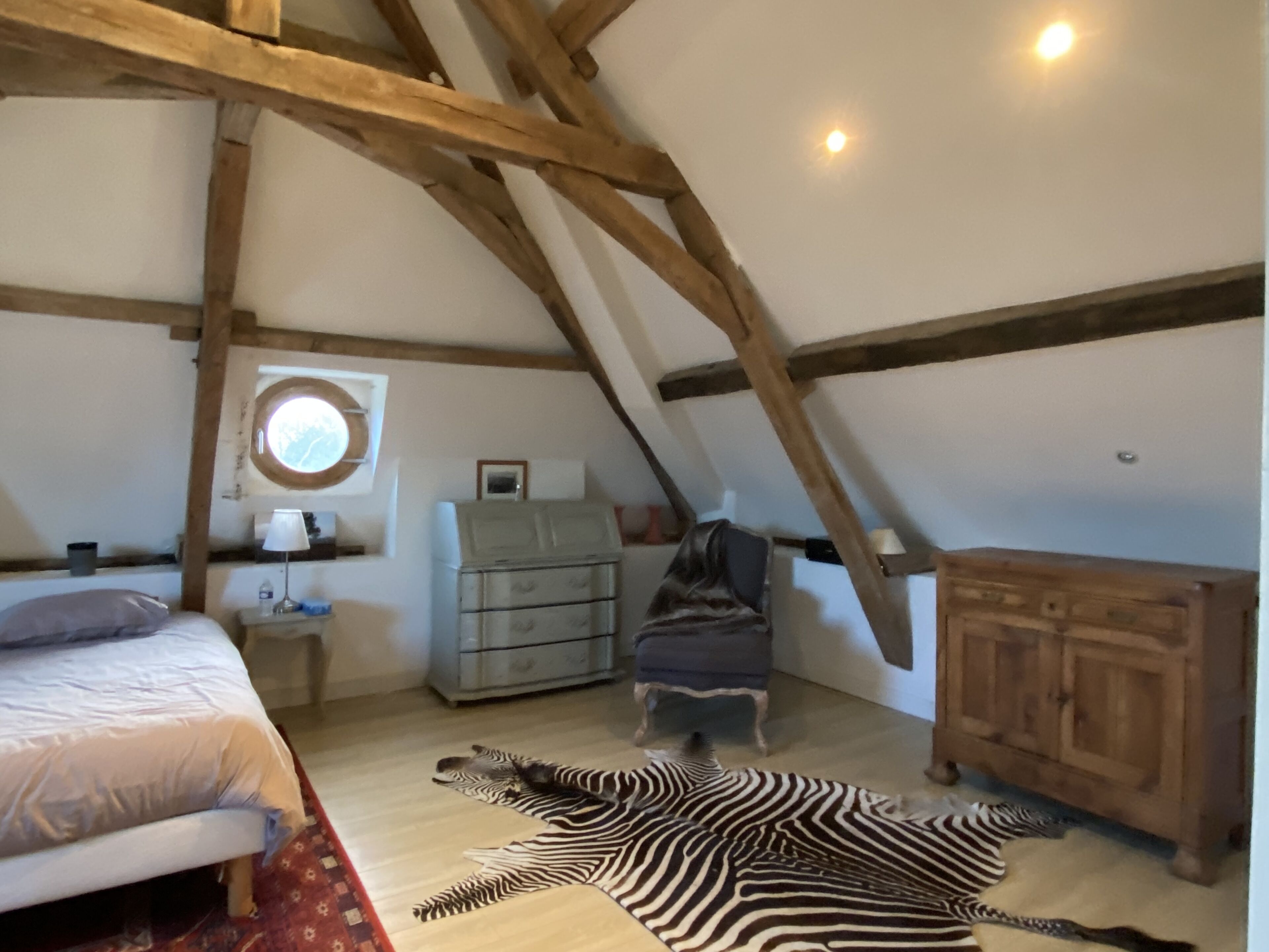 A bedroom with a sloped ceiling, wooden beams, a bed, a dresser, a chair, and a zebra rug.