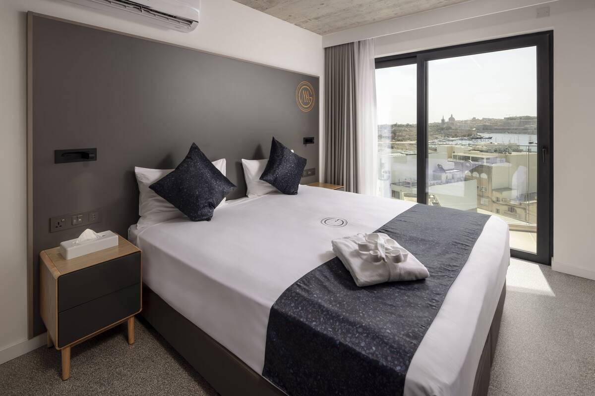 A modern hotel room with a large bed, a nightstand, a window with a city view, and a wall-mounted air conditioning unit.