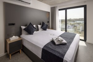 A modern hotel room with a large bed, a nightstand, a window with a city view, and a wall-mounted air conditioning unit.