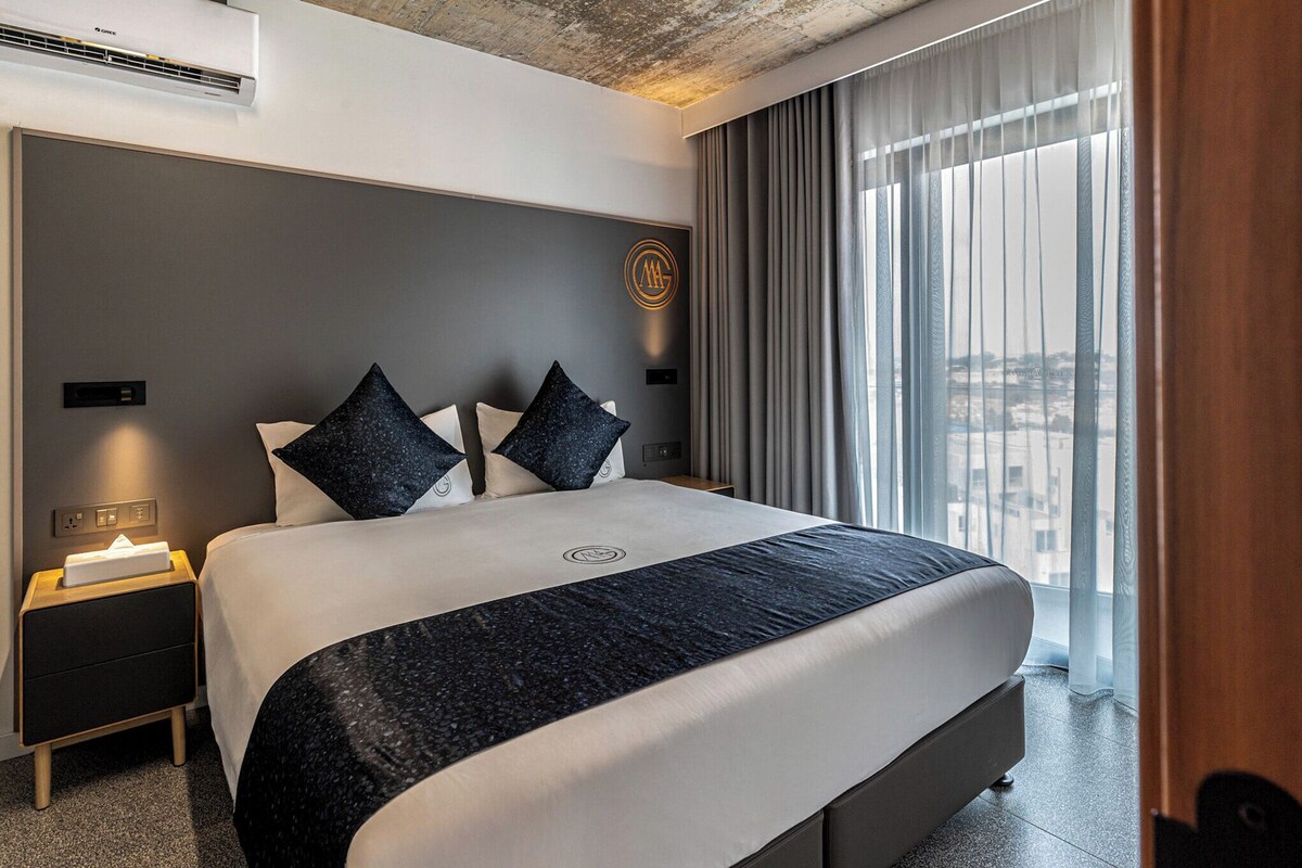 A modern hotel room with a large bed, a nightstand, a wall-mounted air conditioner, and a view through sheer curtains.