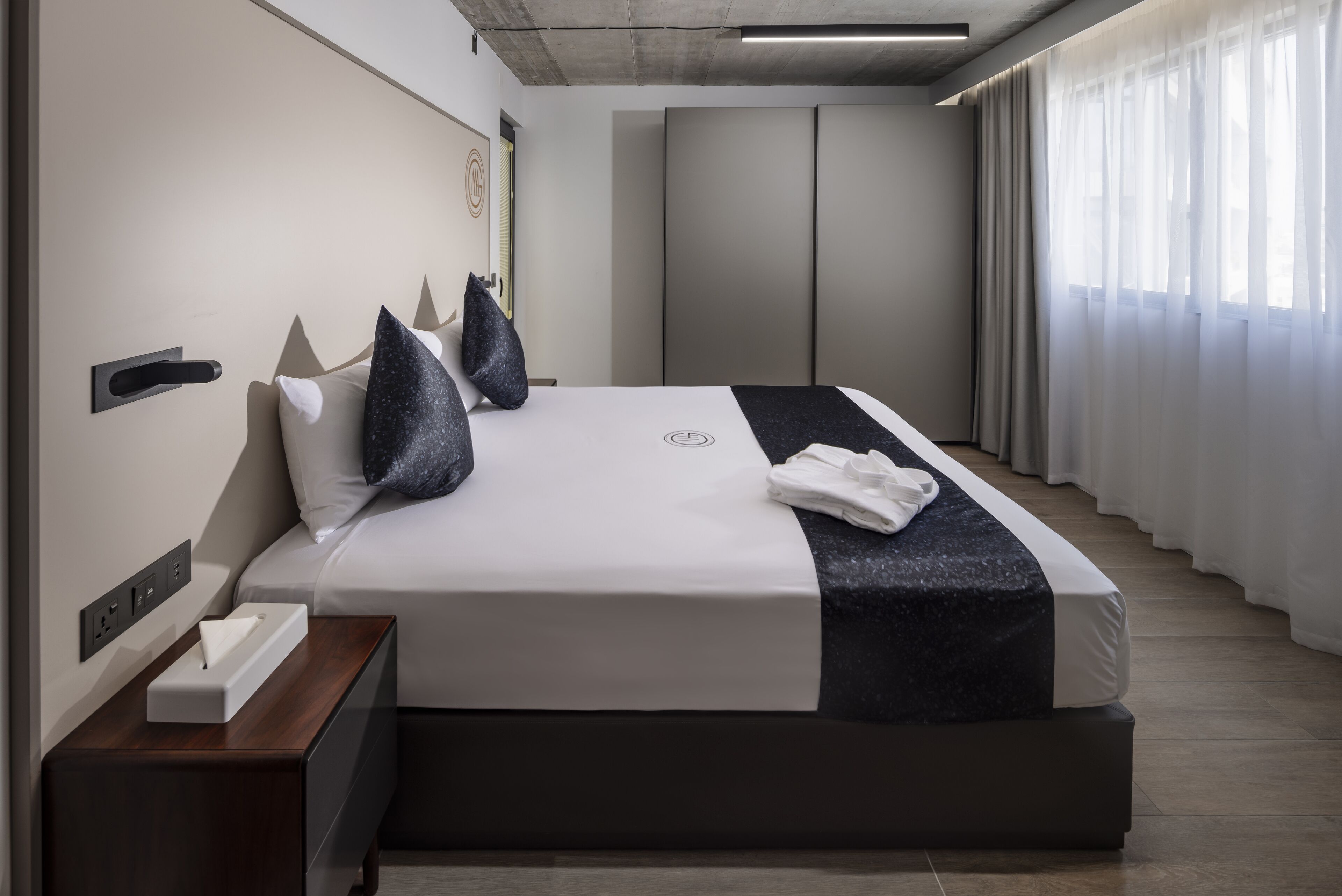 A modern hotel room with a large bed, a bedside table with a tissue box, a wall-mounted light, and a view of the outside through sheer curtains.