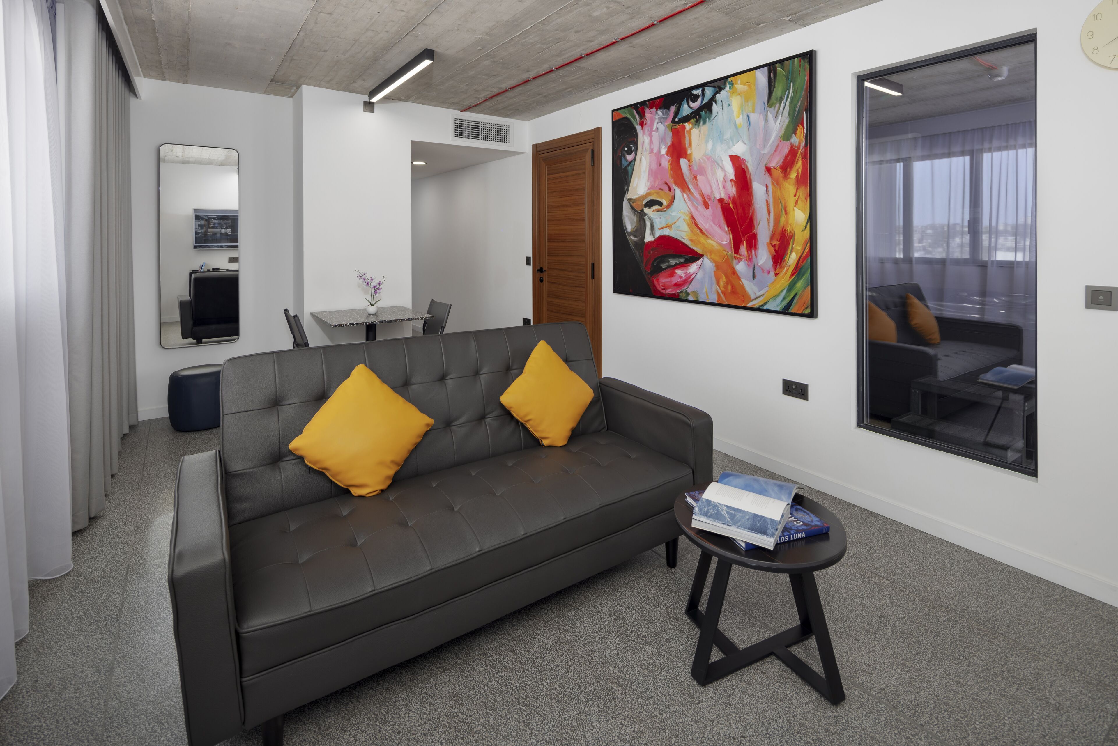 A modern living room with a grey sofa, two yellow cushions, a small round table with a box, a large abstract painting, and a view through a glass door.