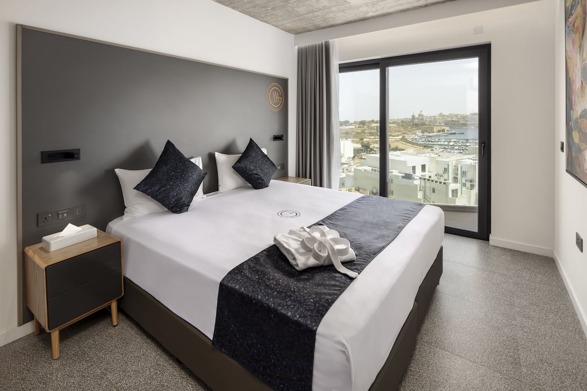 A modern hotel room with a large bed, a nightstand, and a view of the city through a large window.
