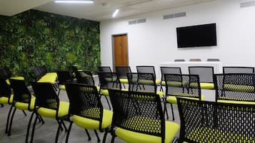 A conference room with rows of black chairs with yellow cushions, a large flat-screen TV on the wall, and a green plant wall.