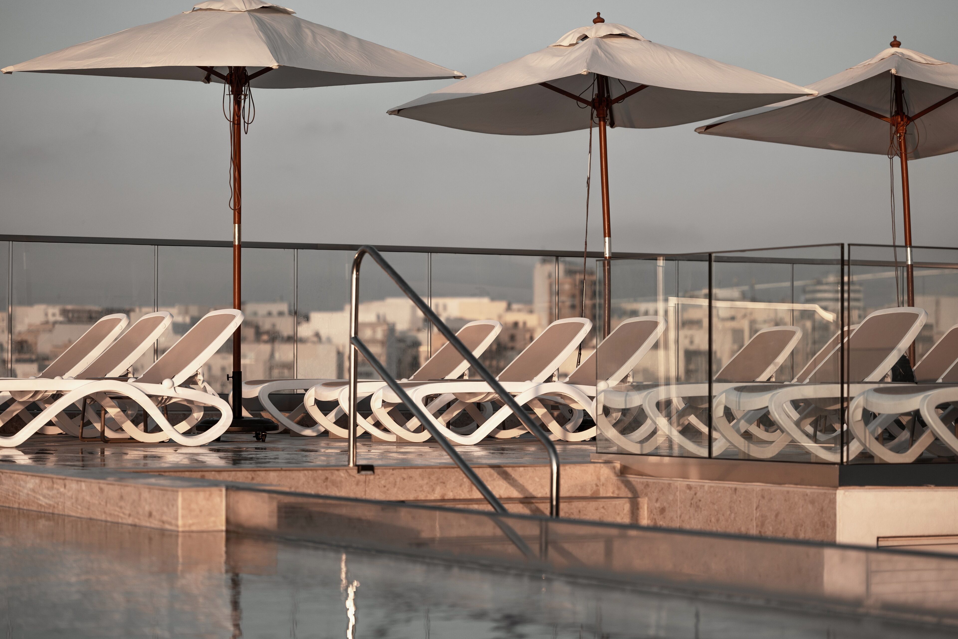 Zona de piscina en la azotea con sillas de descanso y sombrillas, con vistas a la ciudad.
