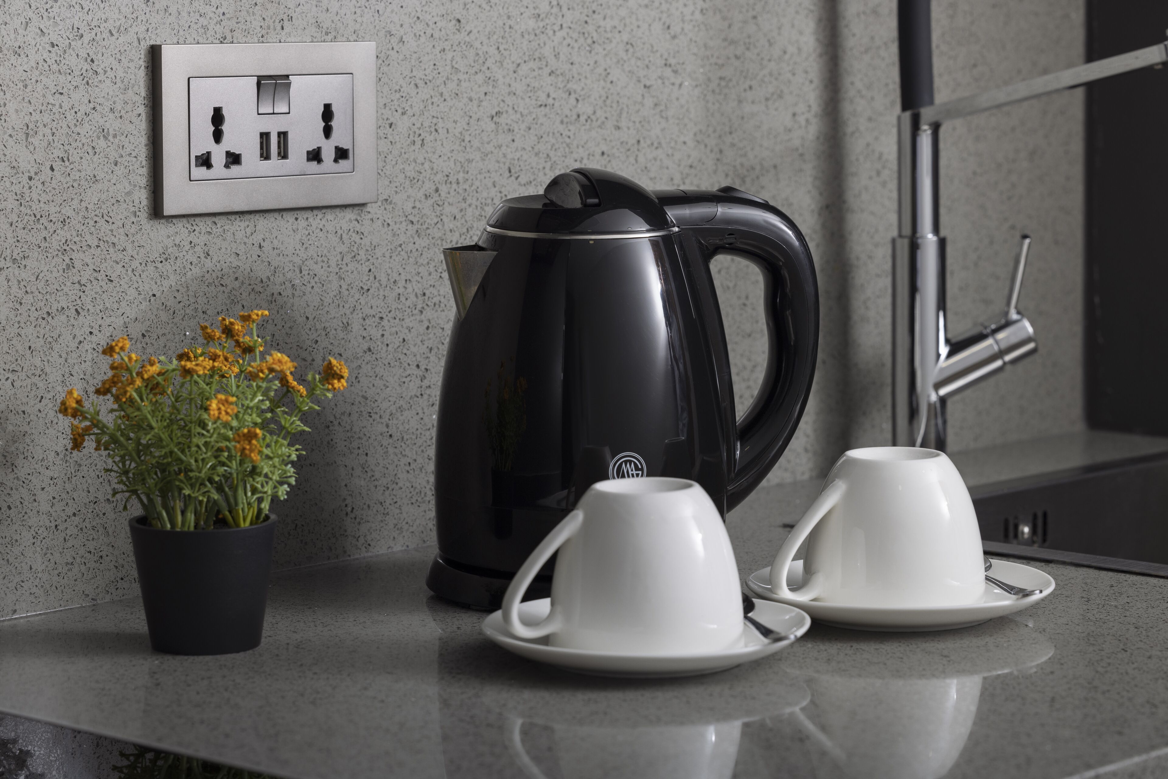 Eine Küchenarbeitsplatte mit einem schwarzen Wasserkocher, zwei weißen Tassen und einem Blumentopf mit gelben Blüten.