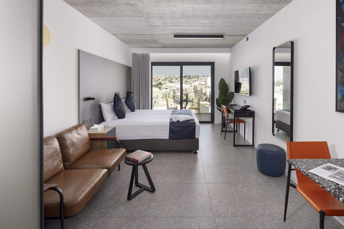 A modern hotel room with a bed, a sofa, a small table, a desk, and a large window offering a city view.