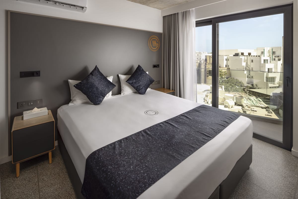 A modern hotel room with a large bed, a nightstand, and a view of the city through large windows.