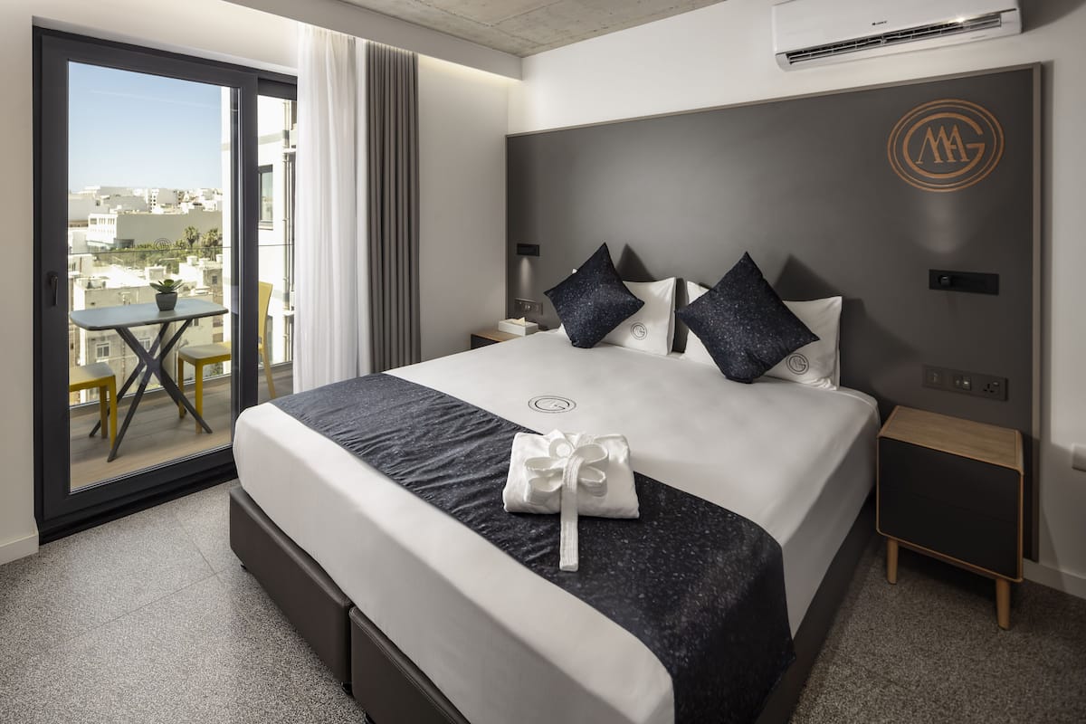 A modern hotel room with a large bed, a small table, and a balcony with a view of the city.