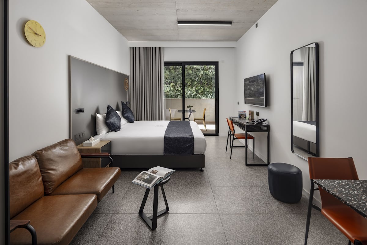 A modern hotel room with a bed, a sofa, a desk, a chair, a small table, a mirror, and a clock on the wall.
