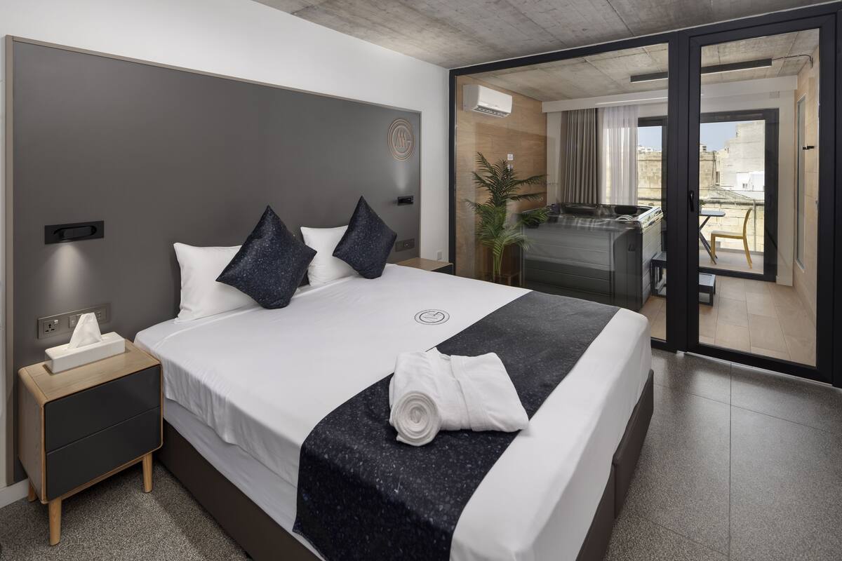 A modern hotel room with a large bed, a bedside table, a small desk, and a view of the city through a glass door.