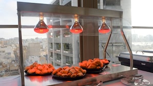 Montaje de bufé con pastelería y una parrilla, visto a través de una ventana con un paisaje urbano de fondo.