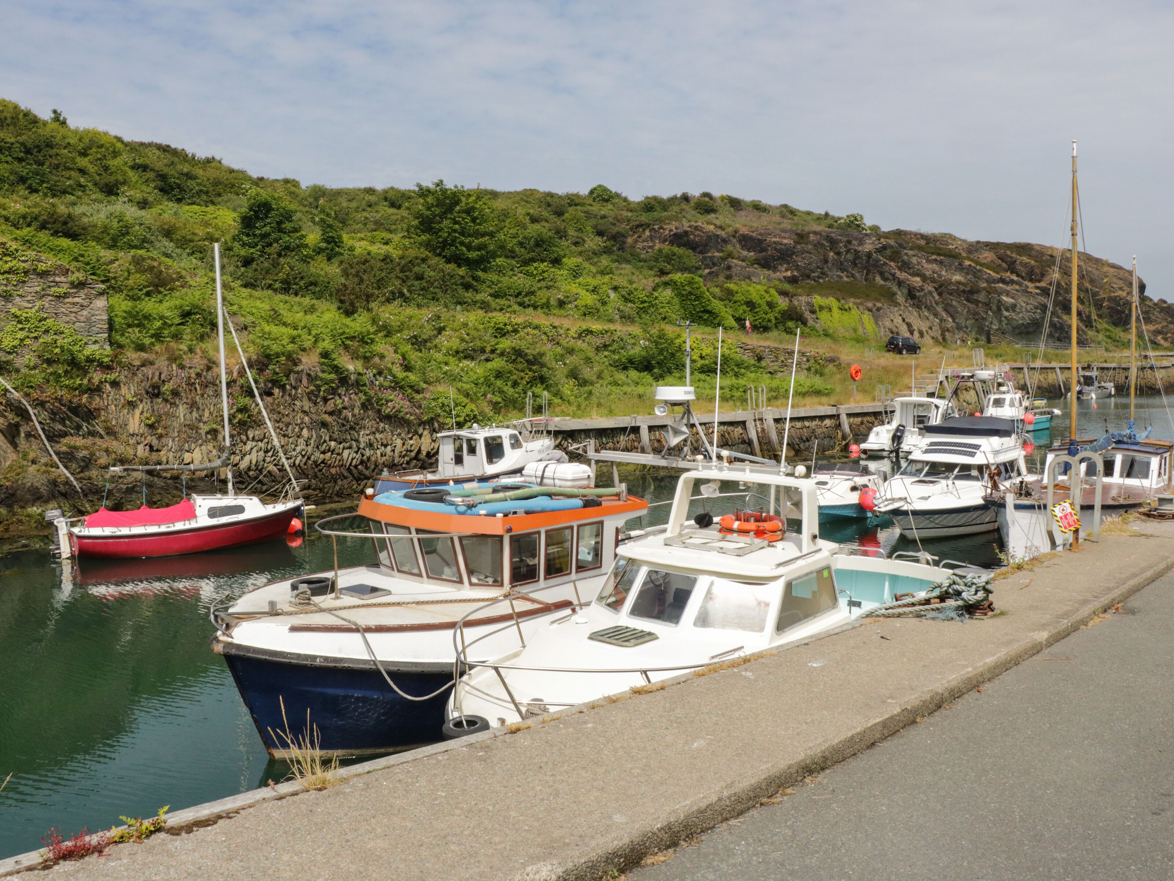 A marina with several boats docked, a rocky hillside in the background, and a concrete walkway along the water.