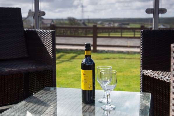 Une table avec une bouteille de vin et deux verres, disposée à l’extérieur, avec des chaises en osier et une vue sur une zone herbeuse et une clôture en bois.