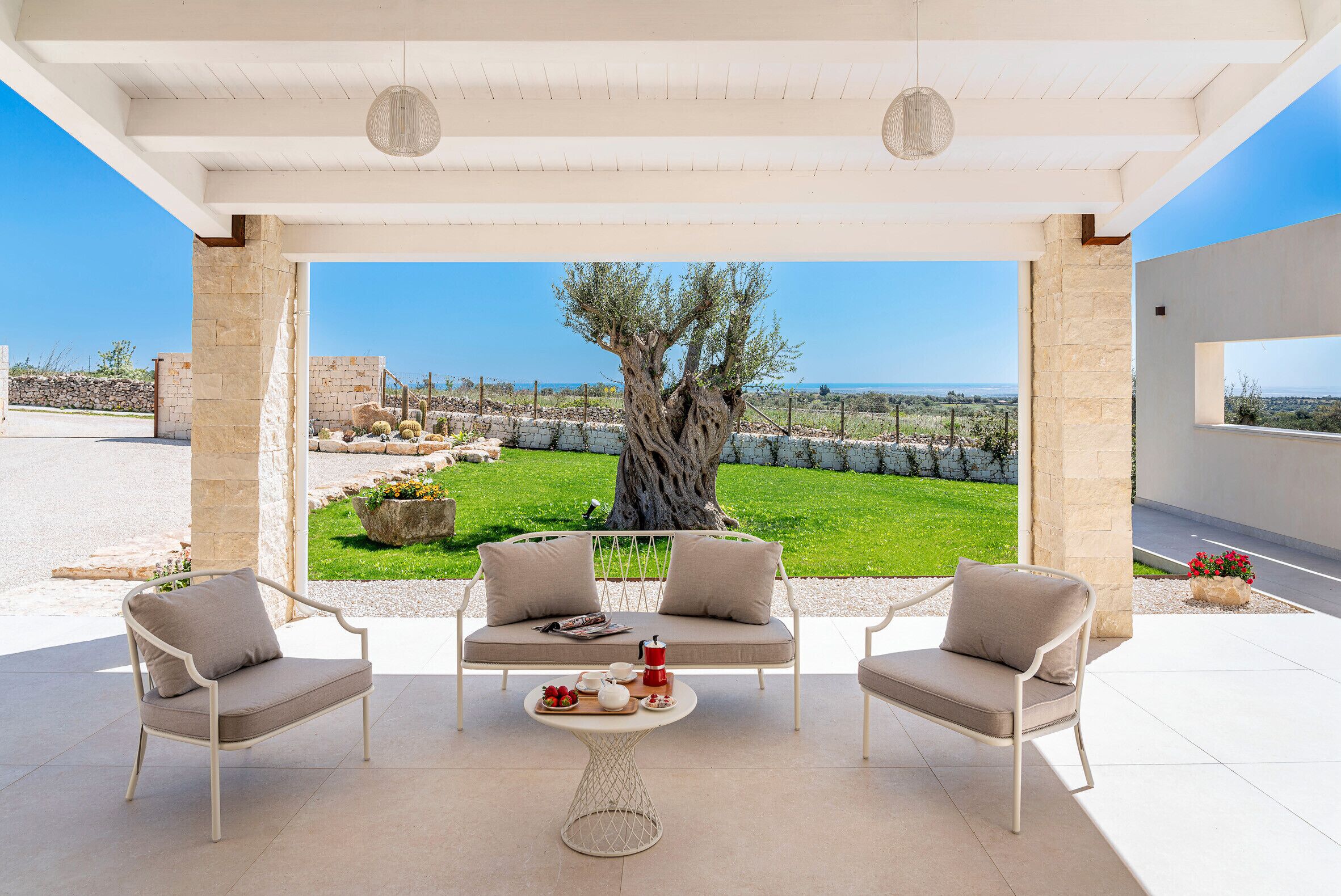 A covered outdoor seating area with a stone table, two chairs, and a sofa facing a view of a tree and landscape.