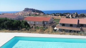 Un quartier résidentiel doté d’une piscine, de maisons aux toits recouverts de tuiles rouges, et, au loin, d’une colline surmontée d’une structure ressemblant à un fort.