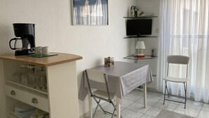 Una habitación pequeña y luminosa con kitchenette, una mesa de comedor con una silla, un televisor en un estante y una ventana con cortinas translúcidas.