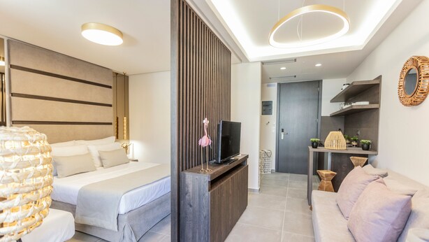 A modern hotel room with a bed, a sofa, a TV on a wooden console, and a small kitchenette area. - Evitel Luxury Living (Kassandra)